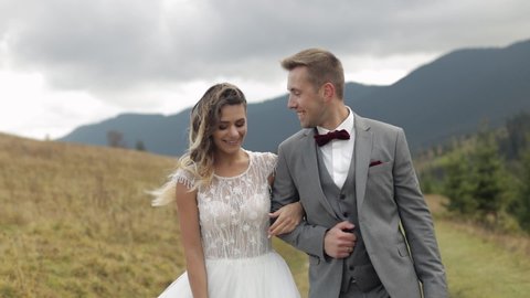 Newlyweds. Lovely young joyful caucasian bride and groom walking on mountain slope hill. Wedding couple. Happy family. Man and woman in love. Autumn tree. Bride in gorgeous wedding dress. Slow motion