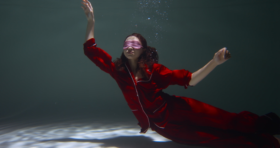 FALLING ASLEEP. Young beautiful woman wearing red pyjamas, sleep mask floats deep under water with open arms slow motion
