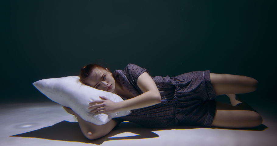 Falling asleep. Cinematic side view of young beautiful woman in pyjamas sinking under water with pillow slow motion.