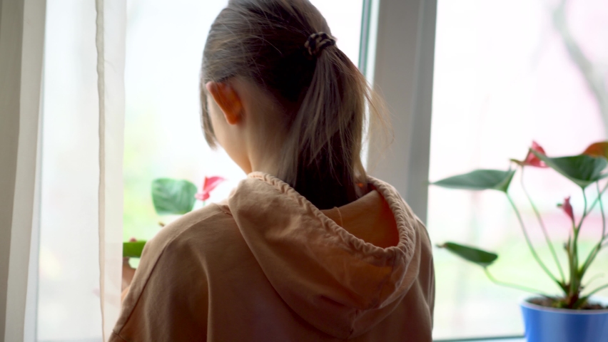 a girl in an orange hoodie looks at a flower by the window and touches it