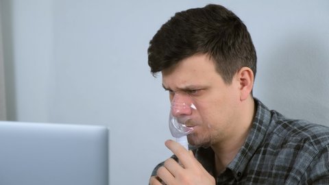 Portrait of sick young man is using nebulizer and inhaler for treatment at home and working on laptop. He is breathing in mask. Fibrosis, covid-19, asthma copd. Treatment, cure and procedure, therapy.