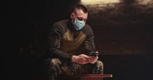 Military man in mask waving hand and speaking while sitting on bench and making video call on smartphone in dark room