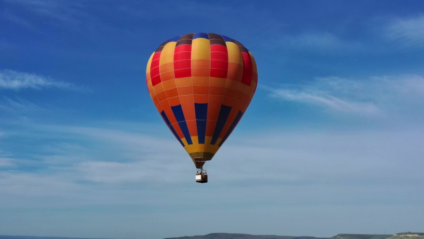 A multicolored hot air balloon with a basket flies against the blue sky. aerial view.  | Shutterstock HD Video #1072103960