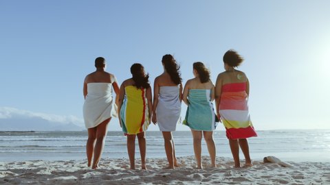 Rear view shot of different sized women wrapped in towel standing on the beach and doing the mexican wave. Multi-ethnic women friends having fun on summer vacation.
