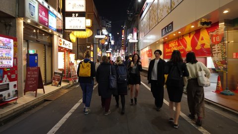 TOKYO - APRIL 03, 2018: Tourist women go at Shibuya central shopping alley at night, first person view camera follow behind. Stores and bright buildings around, many people, night life of Asian city