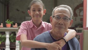 Portrait of Asian adorable young granddaughter embraces her elderly gray hair grandfather happily while posing and looking at camera at home. Togetherness with love in two generations. Healthy life.