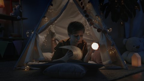 Teen boy in makeshift hut using flashlight while reading book at home in evening. Curious teenager lying on floor while spending free time. Concept of leisure and careless childhood
