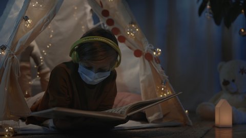 Young boy with headphones spending time in decorative makeshift tent at home in evening. Teen in medical mask lying on floor while reading book. Concept of leisure