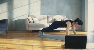 Strong dark haired lady in skinny tracksuit does push ups watching video lesson of training on laptop in living room slow motion