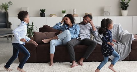 Tired helpless African ethnicity couple sit on sofa feel exhaustion while noisy hyperactive little children running around. Upbringing difficulties, misbehaved son and daughter, ADHD problem concept
