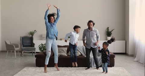 Young african sporty couple with small lively active son and daughter exercising dancing barefoot on carpet warm floor together in modern living room. Happy homeowners family, weekend activity concept