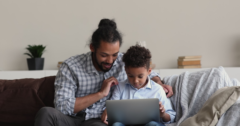 Young African dad and little son sit on sofa using wireless pc, kid and parent buying online, choose video games, learn new app, spend free time on weekend using internet and modern device concept