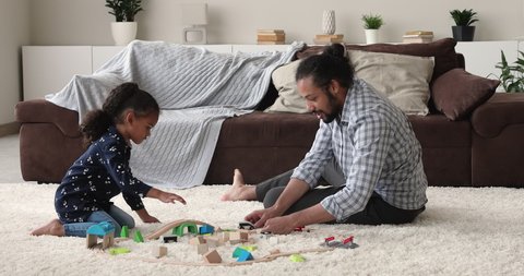 African dad play at home with preschool cute daughter, spend weekend carefree time in modern cozy playroom. Family having fun together enjoy cool railroad wooden toys set sit on floor in living room