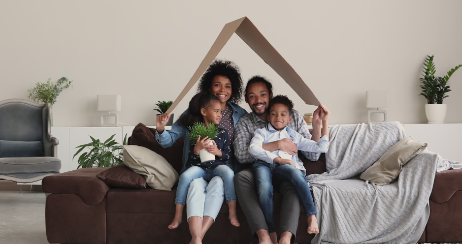 African ethnicity couple with little cheery kids sit under cardboard roof smiling looking at camera. Relocation day to new first home celebration, happy homeowner family portrait, bank loan concept Royalty-Free Stock Footage #1072123643