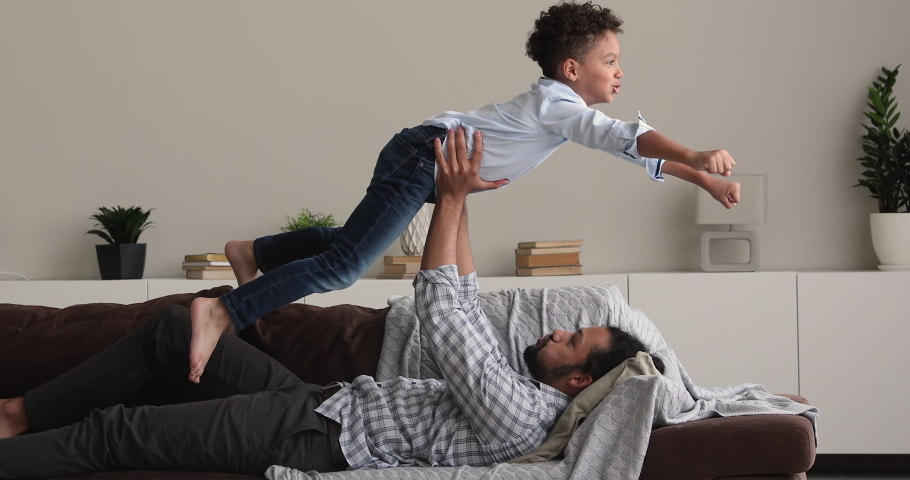 African 6s boy makes gestures with hands imagining like fighting, fly in air while his strong dad lift him on outstretched arms. Father and son playing have fun enjoy creative playtime leisure at home Royalty-Free Stock Footage #1072123658