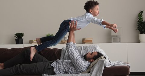 African 6s boy makes gestures with hands imagining like fighting, fly in air while his strong dad lift him on outstretched arms. Father and son playing have fun enjoy creative playtime leisure at home