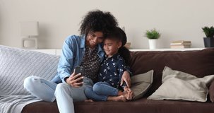 Young African mother little 5s adorable daughter sit on sofa having fun use smartphone, enjoy virtual mobile application, watch videos, taking selfie spend weekend using modern wireless tech concept
