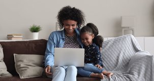Cheerful young African mother and 5s daughter watch cartoon on laptop laughing enjoy carefree weekend together seated on comfy sofa in living room at home. Modern tech, parental control, fun concept