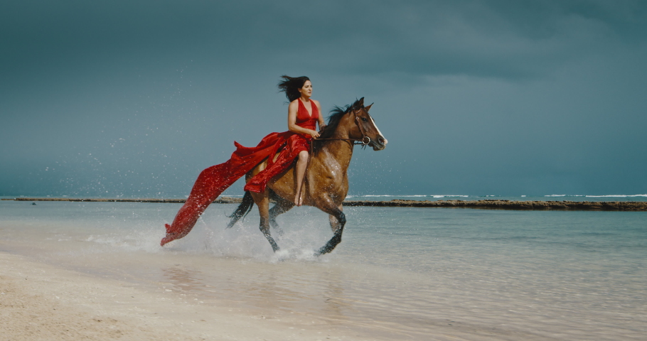 Beautiful woman horseback riding on the beach in flowing red dress, galloping in the water, cinematic slow motion