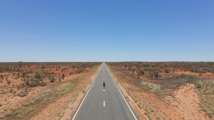 Caucasian Woman Walking On Long And Winding Uluru Road. Australian Outback In The Northern Territory On A Sunny Day. aerial (drone forward)