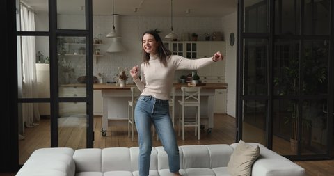 Carefree young vivacious woman dancing alone barefoot on sofa in modern living room at home. Happy female celebrate moving relocation day enjoy new rented apartment. Stress-free weekend, fun concept