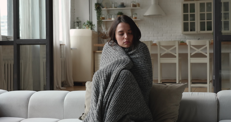 Woman wrapped in warm knitted plaid trembling with cold sit alone on sofa suffers from unbearable cool temperature inside, shivering feels unhealthy. Central heat problems, influenza symptoms concept