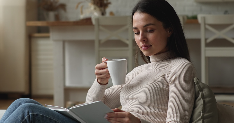 Peaceful millennial woman relaxing seated on comfortable cozy couch hold cup drink tea or morning coffee reading interesting book enjoy favourite literature. Free time, pastime and home hobby concept Royalty-Free Stock Footage #1072143389