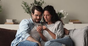 Sincere affectionate young family couple reading email or mobile notification, feeling excited of getting amazing news, celebrating online lottery win together, relaxing on comfortable sofa at home.