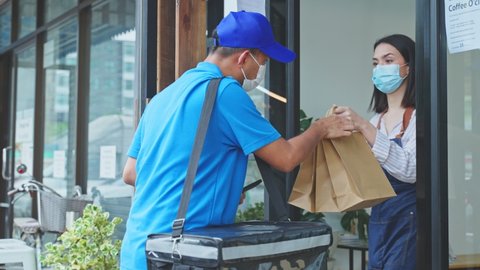 Asian Food delivery man wear protective mask due to Covid-19 pandemic, show online order on phone to restaurant worker at coffee shop. Waitress give takeaway bag to postman for new normal lifestyles.