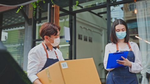 Asian cafe business owner couple wear mask due to Covid-19 pandemic, work in coffee shop. Waiter loads Parcels into car with waitress girl check on clipboard. Delivery Purchased transportation concept