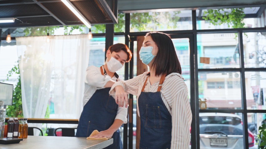 Asian cafe business owner wear protective mask due to Covid19, walk in restaurant with confidence and greeting waiter by elbow bump. Attractive beautiful girl feel happy for new normal in coffee shop. Royalty-Free Stock Footage #1072147718