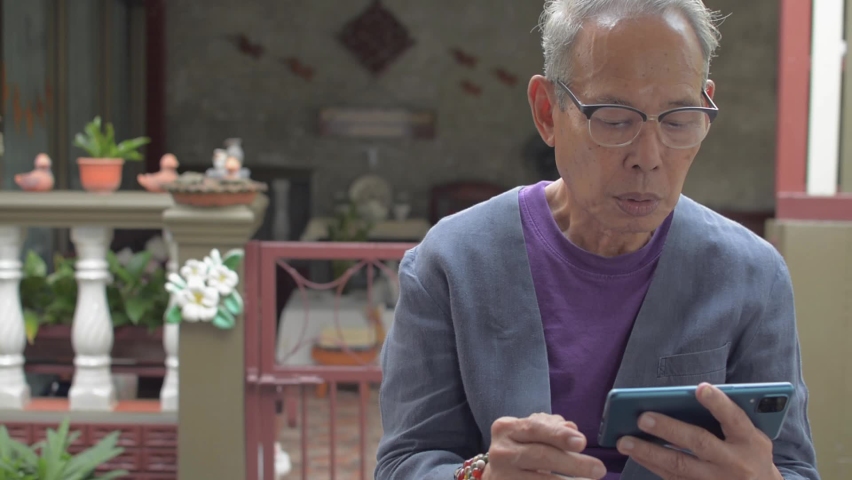 Smiling elderly Asian woman come and hug her gray hair husband from behind while he using and chatting on social network online with mobile smartphone. Senior retired people lifestyle.