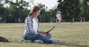 A young student sits on the grass on the territory of the university and communicates via video link on an electronic tablet, is indignant and swears.