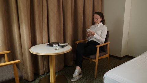 Woman lay aside small notepad and pick up phone, sit resting at chair of hotel room. Business lady have free time to recreate at late evening. She start looking of social networks news feed
