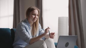 Woman Sitting On Sofa At Home Making Video Call Using Laptop Computer on self isolation. Happy young blonde teacher woman working from home, talking on laptop video conference.