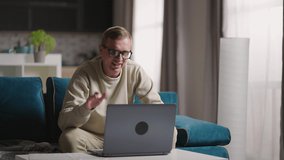 Man Sitting On Sofa At Home Making Video Call Using Laptop Computer on self isolation. Happy young blonde teacher man working from home, talking on laptop video conference.