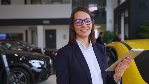 Woman salesperson using tablet and posing for camera while standing in dealership office spbas.