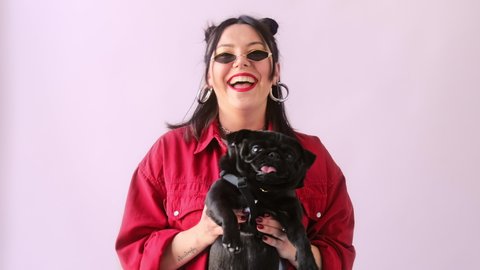 Stylish hipster woman in fashionable glasses dancing and shakes with funny pug puppy in her arms. Dog lover.