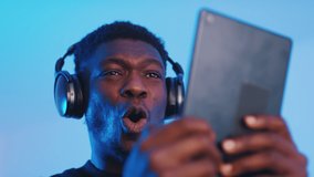 Footage of a guy wearing headphones and holding a tablet watching some video. Intense expressions on his face. Blue background. Closeup of a guy using a tablet watching media. 