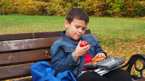 Close-up of a small schoolboy sitting on a Park bench and opening his school backpack during a lunch break. eating a sandwich out of a lunch box
