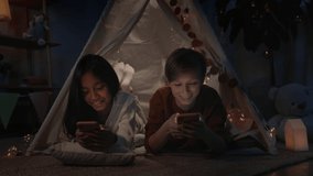 Positive teenagers spending time while watching video in decorative makeshift hut at home in evening. Girl and boy laughing while lying on floor and using smartphones