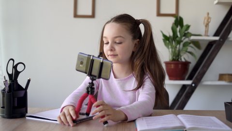 Young caucasian lady authentic puts a tripod on the table with a smartphone for an online lesson in a zoom. The notebook and textbook are open and the child prepares for the video call.