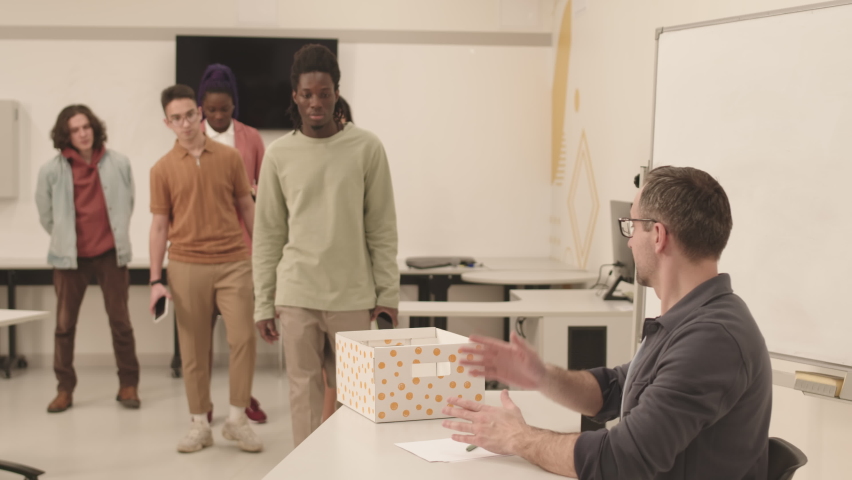 Side view of male Caucasian teacher wearing glasses, sitting at his desk in classroom, pointing hand at box to multiethnic students walking by and handing in smartphones