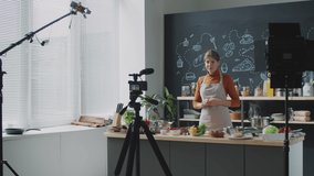 Young woman in apron showing fresh vegetables and talking on camera while filming video for culinary vlog in kitchen