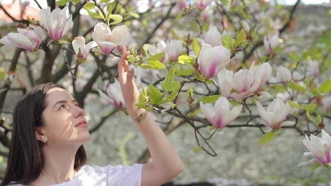 Beauty young woman enjoying nature and touching spring magnolia flowers, Happy Beautiful girl in Garden with blooming magnolia trees. Model smelling blossom, slow motion