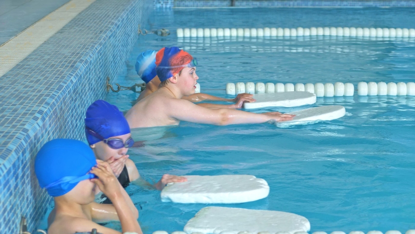Children learn to swim with the help of a swimming board. A swimming instructor teaches children to swim in the pool. Teenagers in swimming suits learn to swim in the pool with an instructor Royalty-Free Stock Footage #1072209452