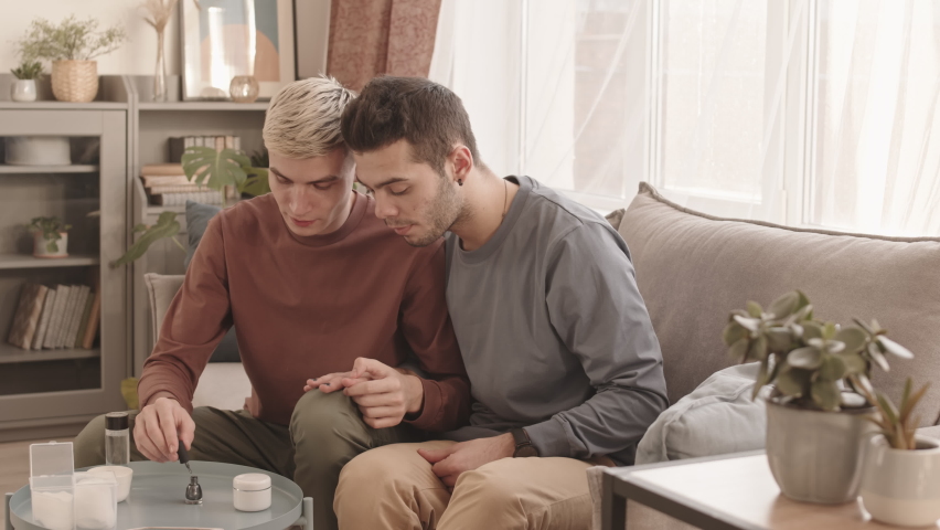 Lockdown of young handsome Caucasian man applying grey nail polish on fingernails of his boyfriend sitting on couch nearby and watching