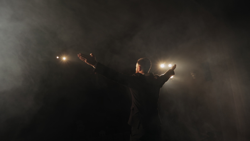 The speaker is standing on stage and holding up his hands. The spotlight is shining on him. There's smoke all around it. 4K Royalty-Free Stock Footage #1072211534