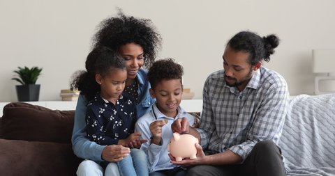 African couple teach little children to save money. Young family with cute kids saving up to joint vacation or new house sit on couch insert coins into piggy bank. Budget management, planning concept