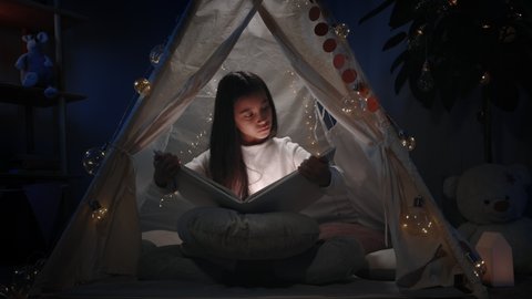 Cute girl with long black hair in decorative makeshift hut looking to book with flashlight. Curious female teenager sitting on floor while reading . Concept of leisure and free time